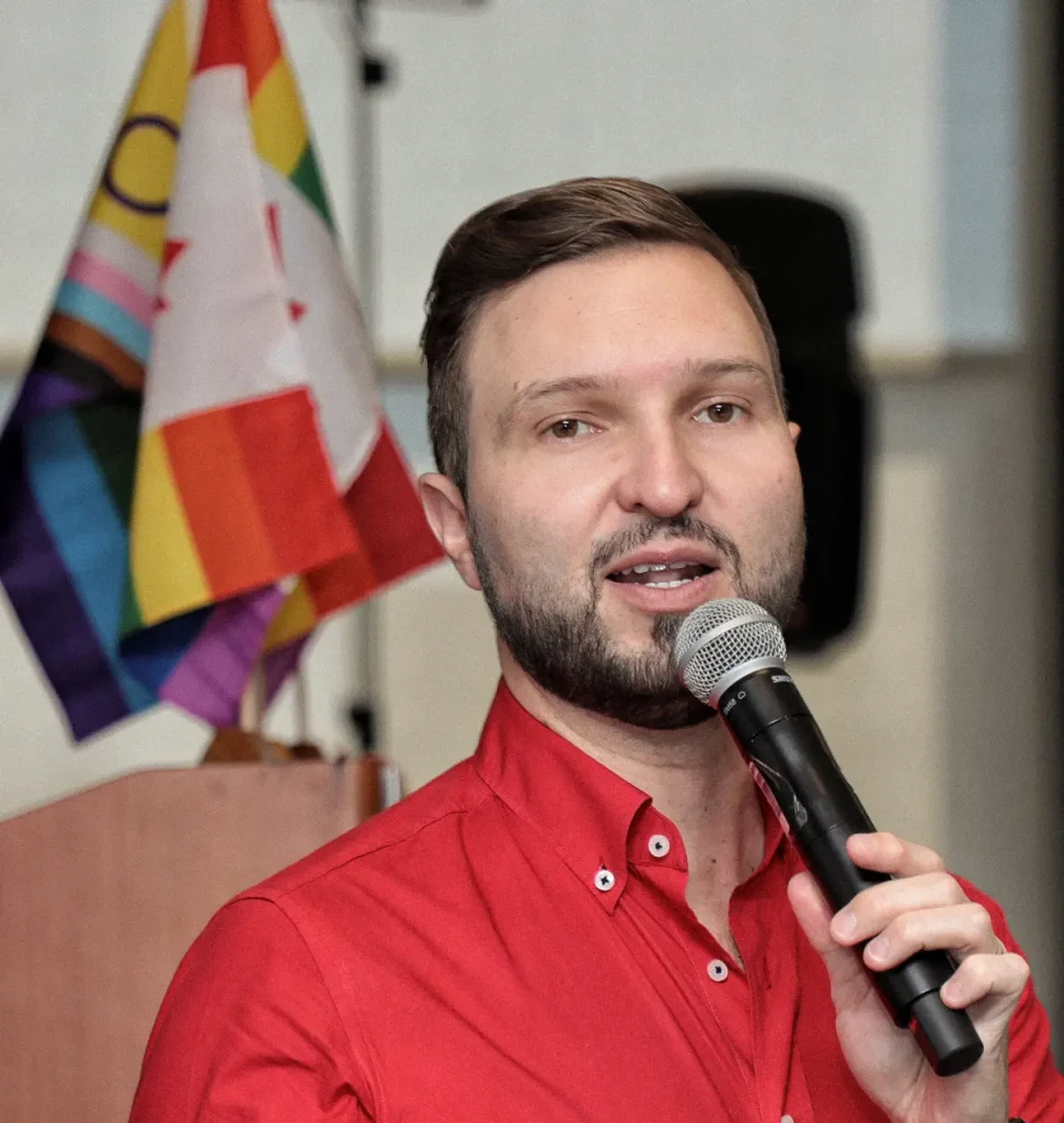 A person in a red button-up shirt speaks into a microphone, with pride flags visible in the background